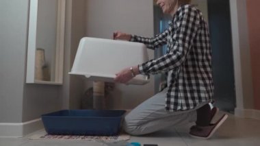 Yaşlı kadın evde evcil hayvanla birlikte yeni kedi kumluğu monte ediyor. Kediyle oynayan yaşlı bir kadın evcil hayvanın tuvalete gitmesi için kapüşonlu çöp kutusu koyuyor. Hayvan bakımı, hijyen konsepti. Kapüşonlu kedi kumu.