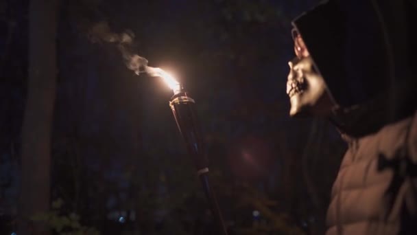 Thème de vacances et fête costumée pour Halloween. Un homme méconnaissable dans un masque de squelette tient une torche de bambou dans sa main et la regarde intensément la nuit. La mort avec une faux et un bâton avec le feu — Video