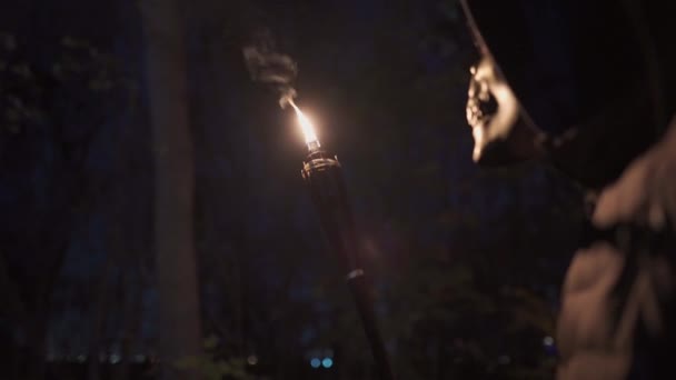 Um homem disfarçado com um esqueleto ou máscara de morte segura uma tocha tiki em sua mão e olha fascinado com o fogo em uma festa de Halloween à noite. Tochas de bambu. Pessoa irreconhecível. Pau queima fogo — Vídeo de Stock