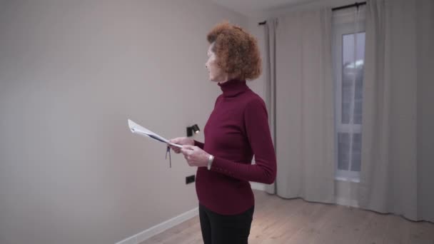 Mulher madura caucasiana a inspeccionar um apartamento vazio antes de assinar um contrato de arrendamento. Uma mulher idosa olha para um apartamento antes de comprar e receber um contrato. Clientes que controlam apartmnet — Vídeo de Stock