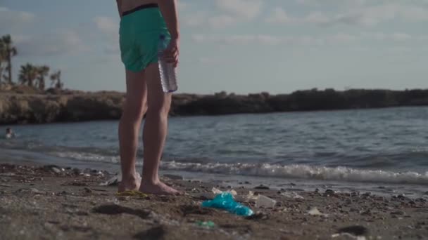 Das Thema Meeresverschmutzung durch Plastik. Ein unkenntlich gemachter Mann steht in einem Müllhaufen an einem Strand in der Nähe des Mittelmeeres und hält eine Plastikflasche in den Händen, die mit dem Rücken zum Horizont blickt. — Stockvideo