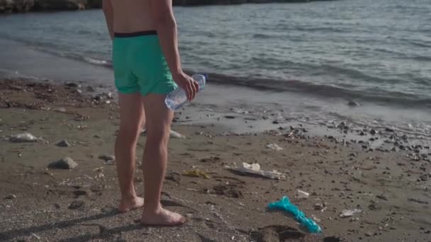 Indiferencia humana a la contaminación de desechos plásticos del mar y del planeta. Hombre irreconocible se para en un montón de basura en la playa cerca del mar mediterráneo sosteniendo una botella de plástico en sus manos mirando al horizonte — Vídeos de Stock