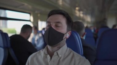 Siyah koruyucu maskeli maskeli maskeli beyaz adam trende şekerleme yapıyor kablosuz küçük beyaz kulaklıklarla müzik dinliyor. Kulaklıklı sıradan bir adam uzun tren yolculuğunda biraz uyuyor.