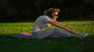 Kafkas formundaki olgun bir kadın parkta ısınmak ve kaslarını yoga minderine germek için egzersiz yapıyor, kulaklıkla müzik dinliyor. Emeklilikte sağlıklı yaşam biçimi kavramı. Yaşlılarda spor