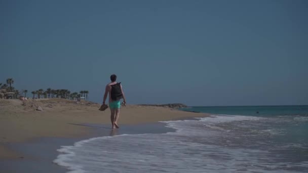 Человек турист с рюкзаком прогуливается босиком по песчаному пляжу возле Средиземноморья Се сзади. Отдыхающий с обувью в руках на пути к месту отдыха на пляже в Пафосе, Кипр — стоковое видео