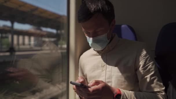 Белый мужчина путешествует в вагоне, сидя в маске и используя смартфон, во время пандемии коронавируса. Тема путешествий и технологий. — стоковое видео