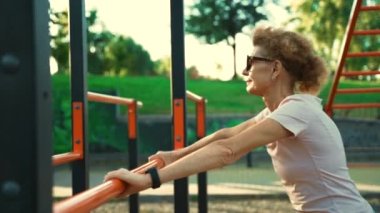 Neşeli, aktif, olgun bir bayan vücudunun üst kısmını çalıştırıyor. Spor salonunun karşısındaki bardan şınav çekiyor.