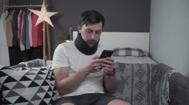 Evde araba kazası geçiren genç adam. Köpüklü tasmalı bir erkek cep telefonundan mesaj okuyor, boynundaki ağrıyı hissediyor. Evdeki kanepede boynu desteklemek için boyunluk takan adam.