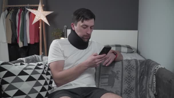 Caucasian male has neck injury to relieve pain, puts on cervical brace made of soft black cotton, male uses smartphone at home sitting on couch in cervical collar, cervical brace relieves tension — Stock Video