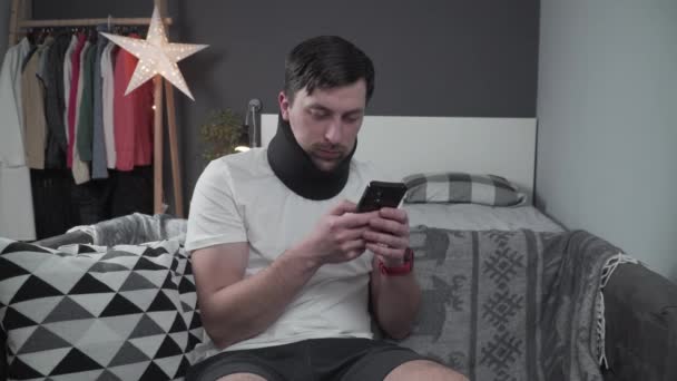 Young man after car accident suffering at home. Male in foam cervical collar reading message on cellphone, feeling pain in neck. Guy wearing neck brace to support neck at home on couch — Stock Video