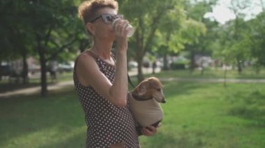 Olgun, narin bir kadın köpeğini bir çanta içinde gezdiriyor ve güneşli yaz havasında kahve içiyor.