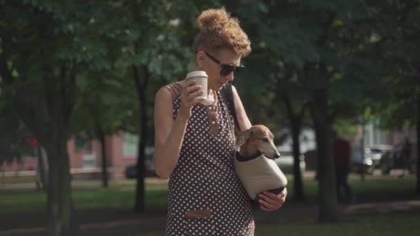 Пожилая кавказка гуляет со своей собакой в мешке для животных и пьет кофе, в солнечную летнюю погоду — стоковое видео