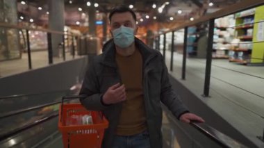 Karantina sırasında konu alışverişi ve market alışverişi, koronavirüs kilitlenmesi. Genç adam alışveriş sepetiyle büyük bir süpermarkette katlar arasında gidip geliyor. Coperid 19 'lu maskeli erkek dükkandan yiyecek alıyor.
