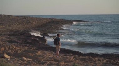 Kıbrıs gezisi teması. Sırt çantalı bir gezgin Paphos şehri yakınlarındaki kayalık bir sahilde Akdeniz 'de gün batımını seyrediyor.