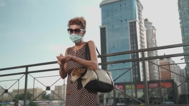 An elderly dog owner in a medical mask uses an antiseptic while walking in the city. Covid time, new rules and restrictions. Mature female in mask disinfecting hands with pet in carrier outside — Stock Video