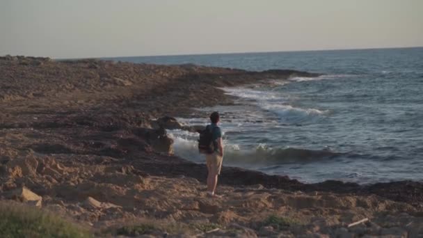 Seorang turis laki-laki dengan ransel mengagumi pemandangan laut Mediterania di pantai berbatu pulau Siprus di kota Paphos saat matahari terbenam — Stok Video