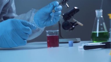 Koruyucu beyaz giysili ve mavi lateks eldivenli bir kimya laboratuarı çalışanının eline yakın plan pipetli kırmızı bir sıvıdan örnek alır.