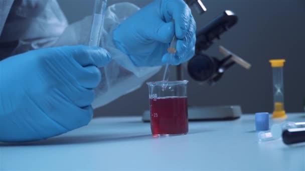 Laboratoire chimique. Un chercheur en vêtements de protection prélève un échantillon de liquide rouge dans une fiole de laboratoire. Gros plan mains. — Video