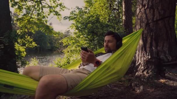 Ao ar livre e conceito de aventura. Viagem de bicicleta para a floresta. Ciclista está descansando em rede verde entre árvores na natureza por lago enquanto ouve música em fones de ouvido e navegar na Internet no smartphone — Vídeo de Stock