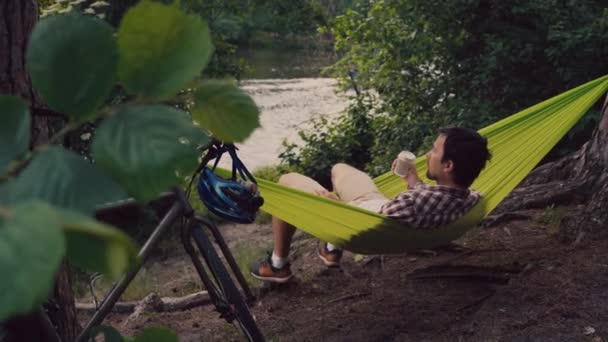 Un hombre llegó al parque en bicicleta y descansa en una hamaca verde con vistas al lago, bebiendo café para ir y disfrutando de la vista de la naturaleza.. — Vídeos de Stock