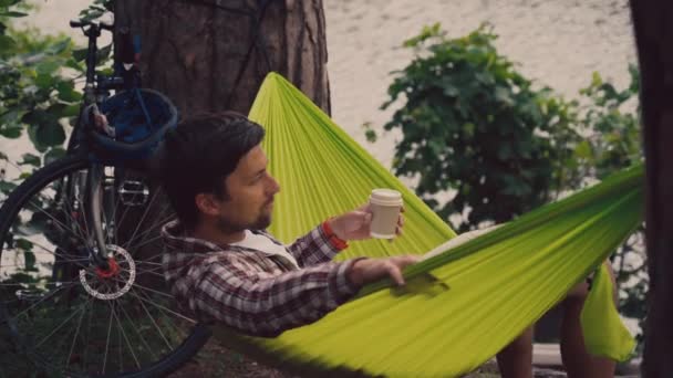 O ciclista balança em uma rede verde e bebe café com vista para o lago na floresta. Um homem descansa em uma rede perto do rio e bebe uma bebida quente depois de andar de bicicleta — Vídeo de Stock