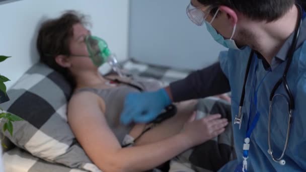 Мужчина в синей форме и женщина в кислородной маске лежат дома на кровати. Врач проводит медицинское обследование женщин, находящихся в карантине с заболеваниями органов дыхания и сердца во время коронавируса — стоковое видео