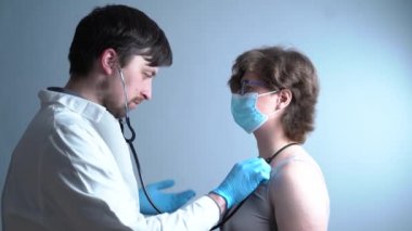 Erkek doktor, gri arka planda tıbbi maskeli bir kadının nefesini kontrol ediyor..