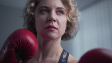 Spor salonunda arka planda gri bir duvarın arkasında kırmızı boks eldivenleriyle poz veren genç ve güzel bir kadın. Sahne arkasında, video çekiminde.