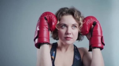 Spor salonunda arka planda gri bir duvarın arkasında kırmızı boks eldivenleriyle poz veren genç ve güzel bir kadın. Sahne arkasında, video çekiminde.