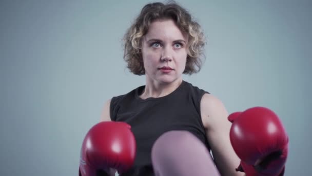 Jeune belle femme mince posant en gants de boxe rouge dans la salle de gym sur le fond d'un mur gris lors d'une séance photo, en coulisses vidéo — Video