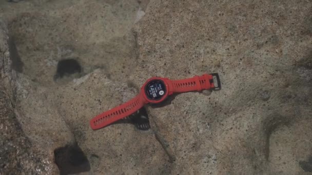 20. května 2021 Kypr, patos. Turistický sportovní hodinky s navigací a červenou srdeční frekvenci monitor garmin instinkt plamen červená v čisté mořské vodě na skále — Stock video