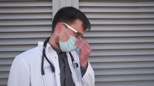 クリニックの外で疲れた若い男性医師の肖像画は 疲労で目をこすり 頭痛を感じ 彼の医療マスクや眼鏡を離陸します 病院で非常に長いシフトの後に休んでいる深刻な医師 — ストック動画