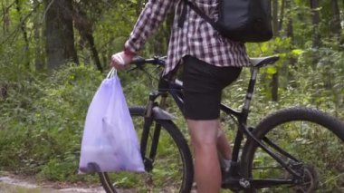 Erkek bir bisikletçi, doğada bisiklet sürerken ormanda plastik çöp topladıktan sonra çöp torbasıyla ayakta duruyor. Çevreyi koru. Çevre koruma, doğada toparlanma. Kirlilik. Ekolojik sorun