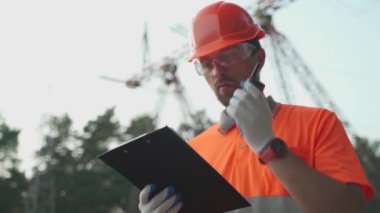 Elektrik santrali işçisi yürüyor ve telsizle rapor veriyor. Açık hava çalışma alanındaki veri tablosunda turuncu koruyucu iş elbiseleriyle elektrik hatlarıyla karşılaştırılıyor. Radyo istasyonunu kullanan mühendis 