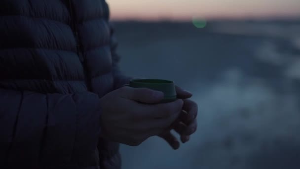 在日落时分 手持一个带着热饮的旅行杯的徒步旅行男性游客 在营地里 把一个穿着绒毛夹克的旅行者的茶杯折叠起来 早上喝着热饮户外厨房 — 图库视频影像