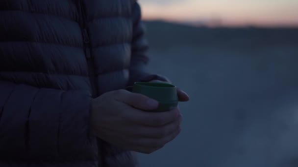 ハイキングキッチン ダウンジャケットの男の旅行者の手が凍り 日没のキャンプ場で熱いキャンプカップに彼の腕を暖めます 折り畳み式のカップの中で熱い飲み物 コーヒーを屋外で 登山家がお茶を飲む — ストック動画