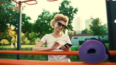Sportif olgun bir kadın yoga dersinden sonra sokak spor salonunda telefon kullanıyor. Üst düzey Kafkasyalı bayan dışarıda yoga antrenmanından sonra mola veriyor. Aktif yaşam tarzı konsepti. İnsanlar, spor ve teknoloji.