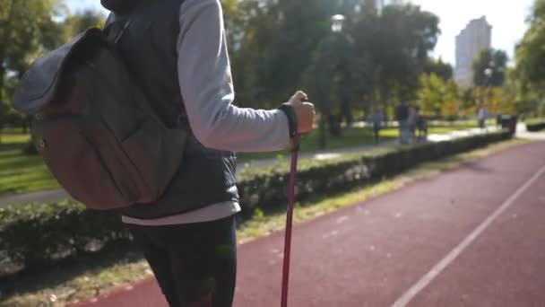 北欧人老年时带着两根柱子散步的主题 老年时的积极休闲和锻炼 在阳光充足的天气里 拿着手杖走路的女人在公园的跑道上走着 斯堪的纳维亚棍棒 — 图库视频影像