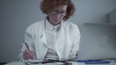 Bayan kıdemli doktor tıbbi raporlar hazırlıyor. Klinikteki dizüstü bilgisayarının başında otururken kalemle tıbbi belgelere not alıyor. Günlük hekimlik işi. Aile doktoru reçete yazar