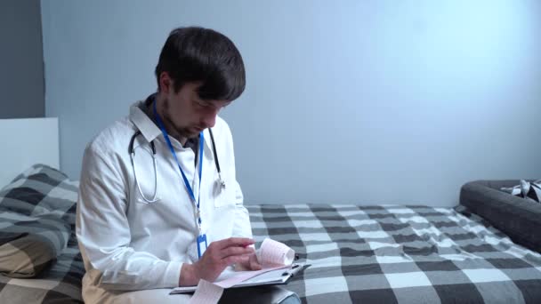一名男医生坐在卧室的床上 检查家中病人的心电图结果 心脏科专家家访同一病人 医生检查心电图 — 图库视频影像