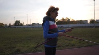 Yaşlı kadın şehir stadyumunda İskandinav yürüyüş direkleri açıyor. Yaşlı bir dişi, İskandinav bastonlarını uzatır ve hazırlar. Olgun sporcu antrenmandan önce İskandinav çubuklarını açıyor