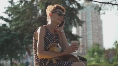 Yetişkin bir evcil hayvan sahibi bankta oturur, kahve içer ve bir yaz parkındaki bankta telefonla konuşur. Yaşlı bir kadın telefonda konuşuyor ve elinde bir köpekle kahve içiyor.