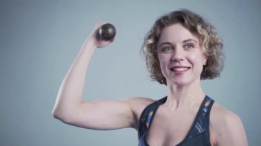 Spor salonunda gri bir duvara karşı poz veren halterli bir kadın. Elinde kettlebell olan atletik bir kadın için sahne arkası fotoğraf çekimi. Gülümseyen sporcu kadın iki halterle egzersiz yapıyor ve poz veriyor.