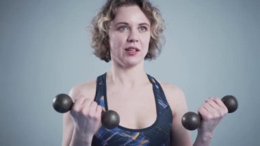 Stüdyodaki bir bayan atlet için dışarıdan çekilmiş bir fotoğraf çekimi. Elinde dambıllarla poz veren zayıf atletik bir kadın. Spor kıyafetleri ve dambıllar fotoğraf için poz veriyor.