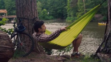 Genç beyaz bir adam gölün yanındaki ormanda bisikletini sürdükten sonra hamakta dinleniyor. Cennet gibi bir yer. Seyahat, doğada kamp. Hippi bisikletçi nehir kenarındaki bir hamakta eğleniyor.