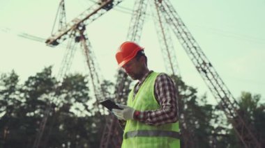 Enerji endüstrisi mühendisi elektrik santralinde koruyucu yelek ve başlık takan tabletle çalışıyor. Mühendis tablet kullanarak yüksek voltaj güç şebekesini inceliyor. Arka planda elektrik direkleri ve elektrik hatları