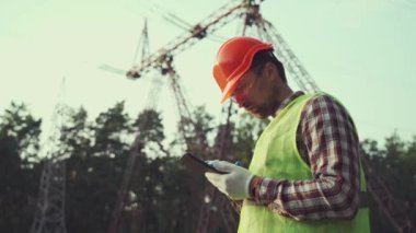 Enerji endüstrisi mühendisi elektrik santralinde koruyucu yelek ve başlık takan tabletle çalışıyor. Mühendis tablet kullanarak yüksek voltaj güç şebekesini inceliyor. Arka planda elektrik direkleri ve elektrik hatları