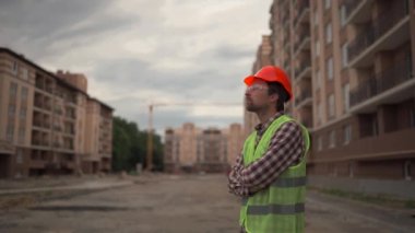 Thoughtful male construction worker thinks about something with arms folded across his at construction site. Handyman in reflective vest, helmet, and safety glasses with his hands on chest proudly