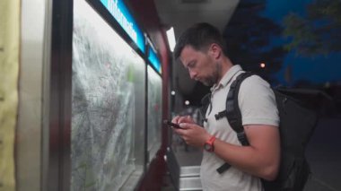 Male passenger analyzes transport card at a public transportation stop and matches it to an app on his phone in Munich, Germany. Checking map and waiting for transport in city. Public transport route