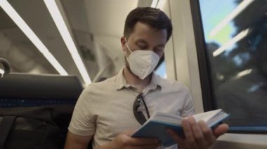 Passenger on public transport is reading a book while sitting by window wearing a protective mask. A man traveling on a Deutsche Trains wearing a protective mask and reading a book. German railroad. 